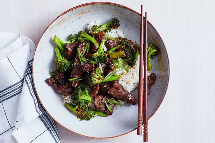 A bowl of beef and broccoli stir-fry served over white rice, garnished with sliced green onions, with chopsticks resting on the bowl and a white napkin nearby.
