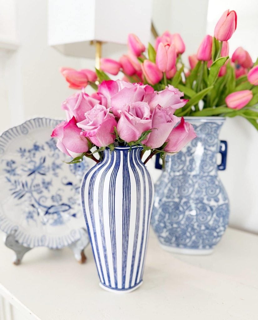 A blue and white striped vase with pink roses is displayed in front of a blue floral plate and a blue-patterned vase with pink tulips.