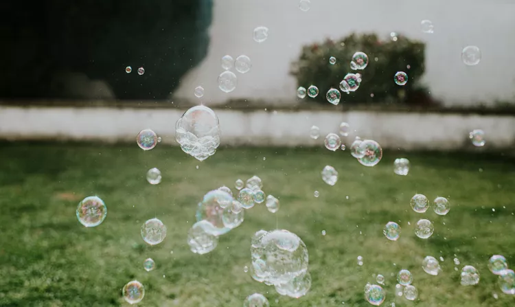 Several soap bubbles floating in the air above a grassy lawn with a blurred white wall and shrubbery in the background.
