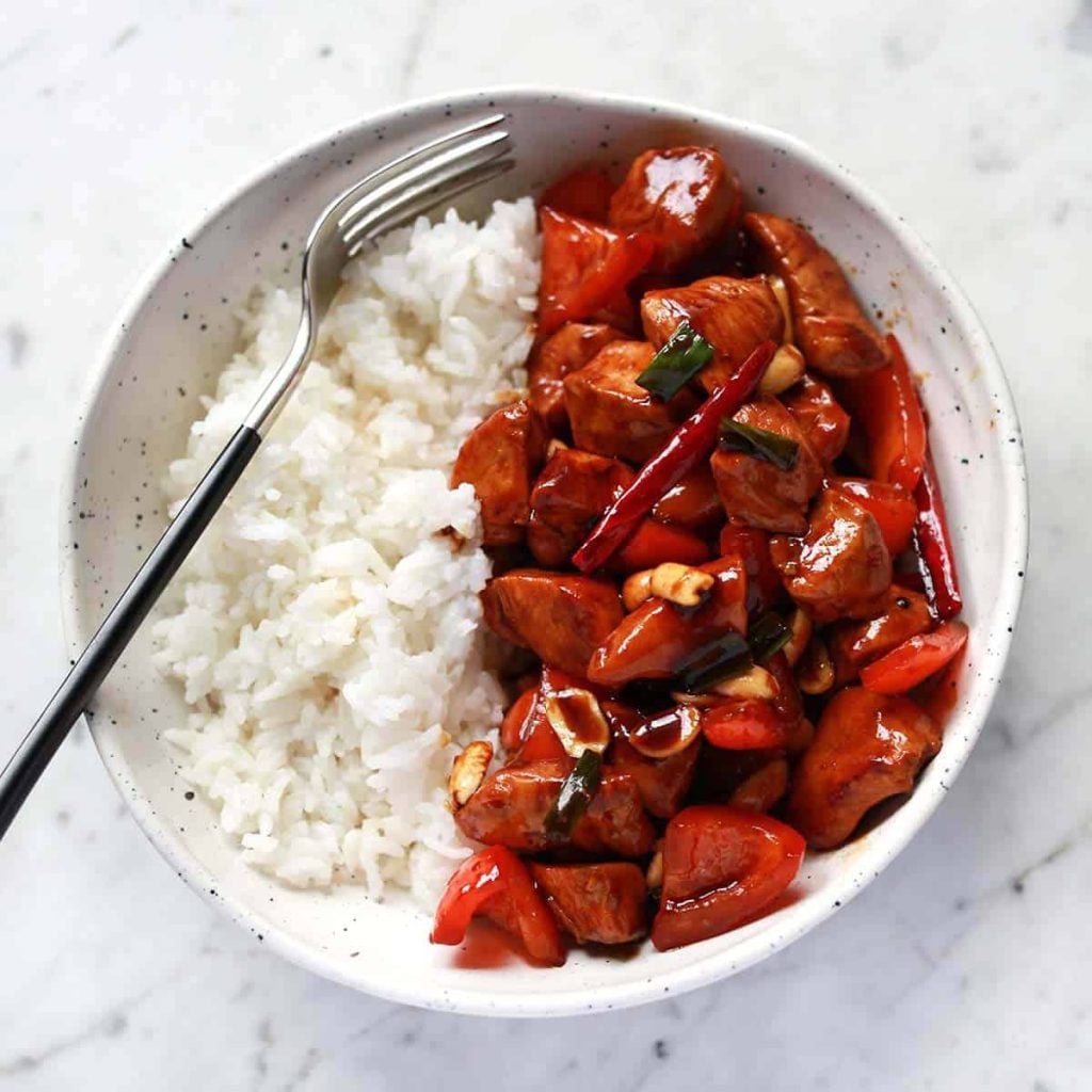 A bowl of white rice and stir-fried chicken with vegetables and sauce, served with a fork on a white marble surface.