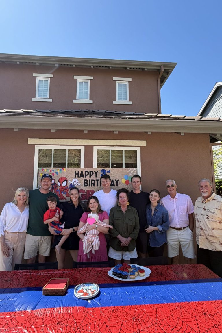 A group of people pose for a birthday photo outside, standing in front of a house with a decorated window and a table set with cake and treats.