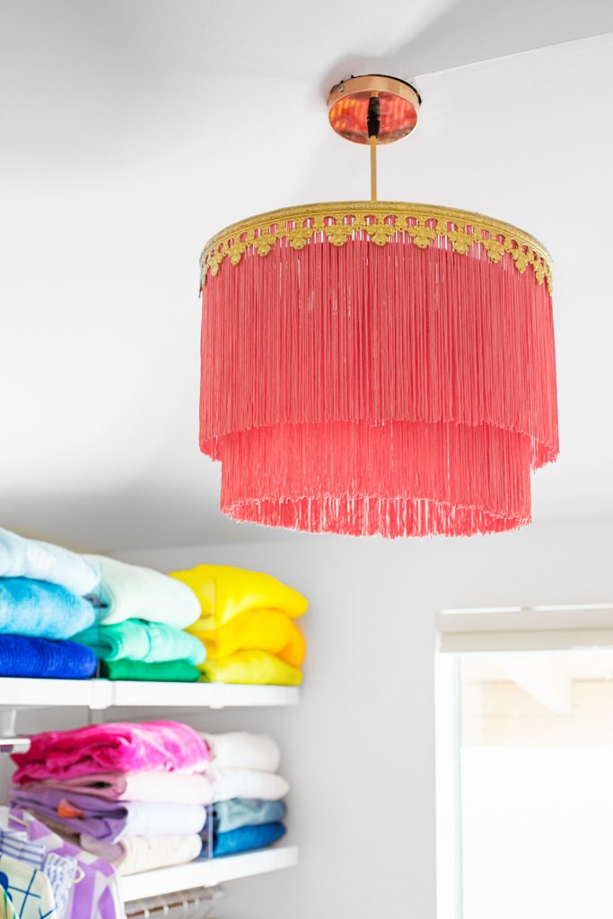 A gold and pink fringe chandelier hangs from the ceiling above neatly folded, colorful towels on white shelves.