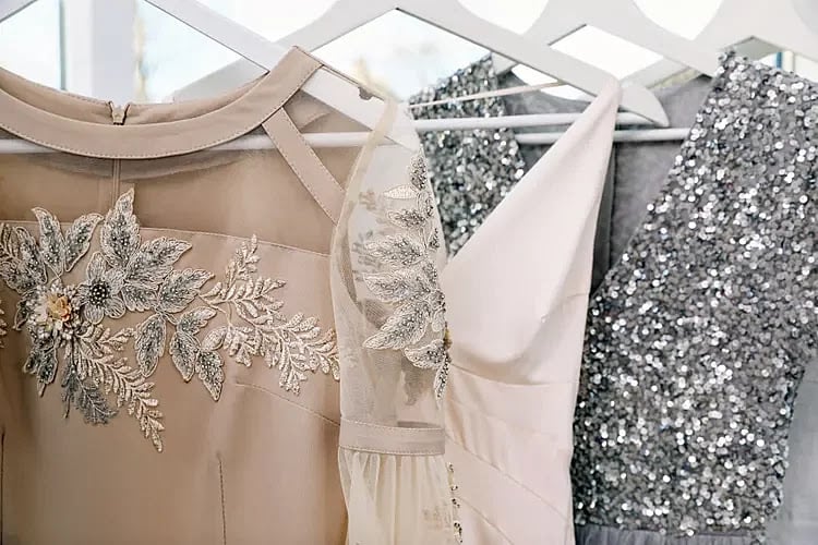 Three formal dresses hanging on white hangers: one beige with floral embroidery, one plain light-colored, and one gray with silver sequins.