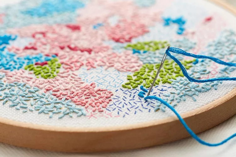 Close-up of a hand embroidery hoop with a threaded needle stitching a colorful pattern using blue, pink, and green threads.