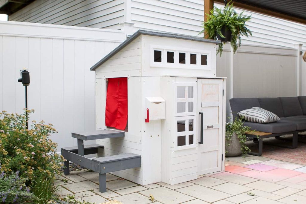A small white wooden playhouse with gray steps, a red curtain, and a mailbox, situated on a patio near outdoor seating and plants.