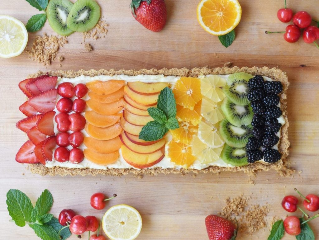 Rectangular fruit tart with rows of strawberries, cherries, apricots, oranges, kiwi, and blackberries on a wooden surface with scattered fruits and mint leaves.