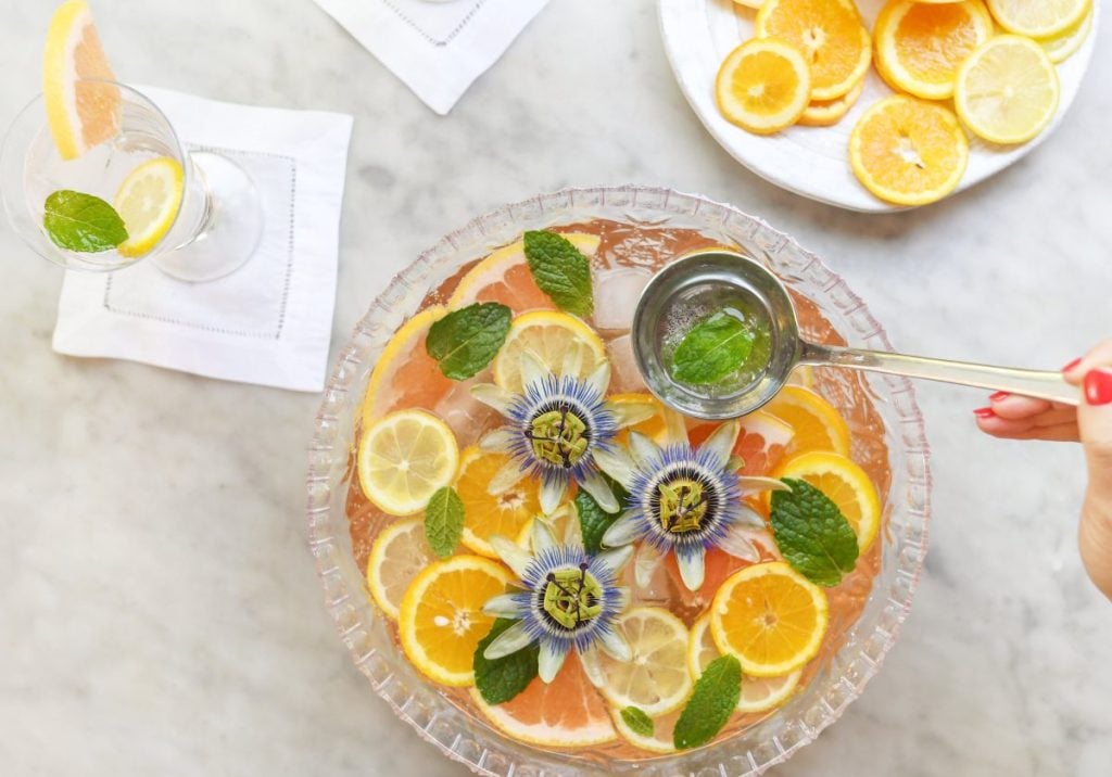 A glass punch bowl with lemon and orange slices, mint leaves, and purple flowers, next to a glass with a mint garnish and a plate of citrus slices. A hand ladles punch into the glass.