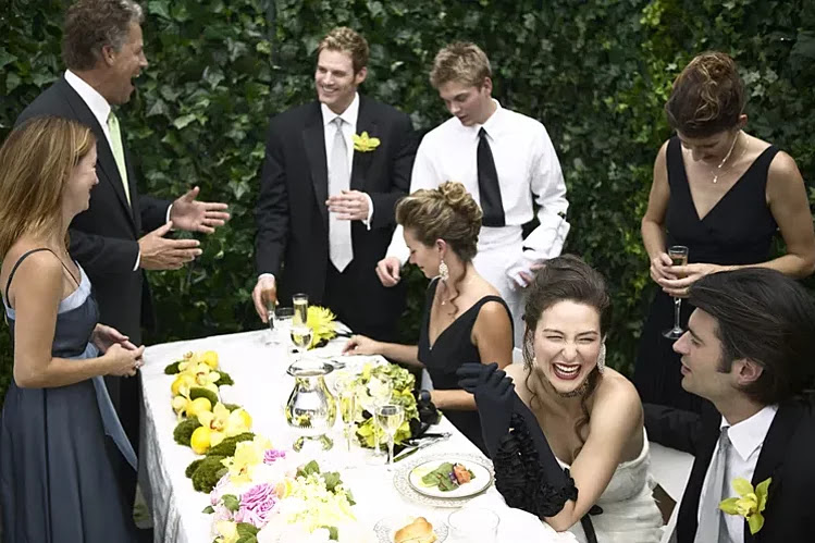 A group of people dressed formally are gathered around an outdoor table with food and drinks, laughing and engaging in conversation.