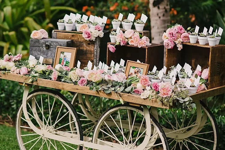 A decorative cart with wooden crates holds framed photos, small potted plants with name cards, and pink flower arrangements, set outdoors with greenery in the background.