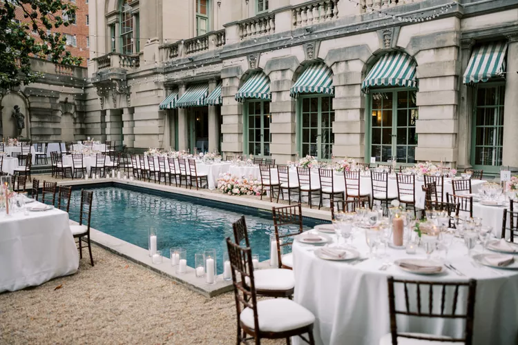 Outdoor wedding reception setup by a rectangular pool with round and long banquet tables, white linens, and floral centerpieces, adjacent to a historic building with striped awnings.