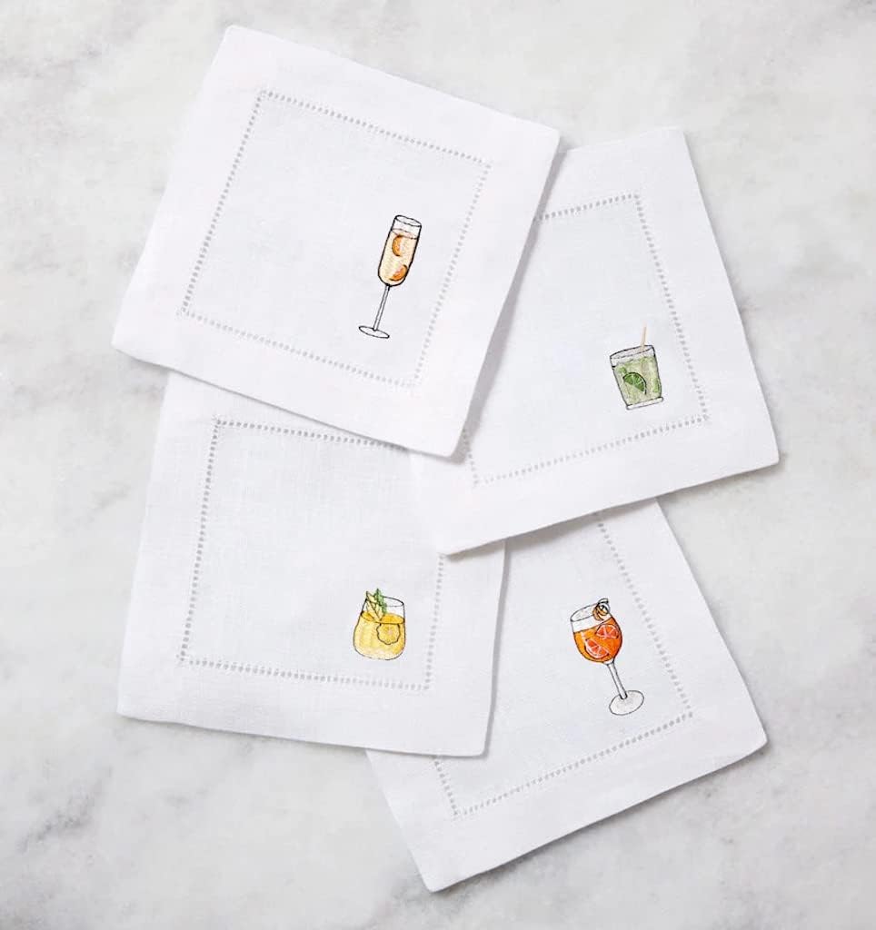 Four embroidered cocktail napkins in crisp white linen, each adorned with a unique drink design, are artfully arranged on a marble surface&mdash;perfect for elevating your next gathering.