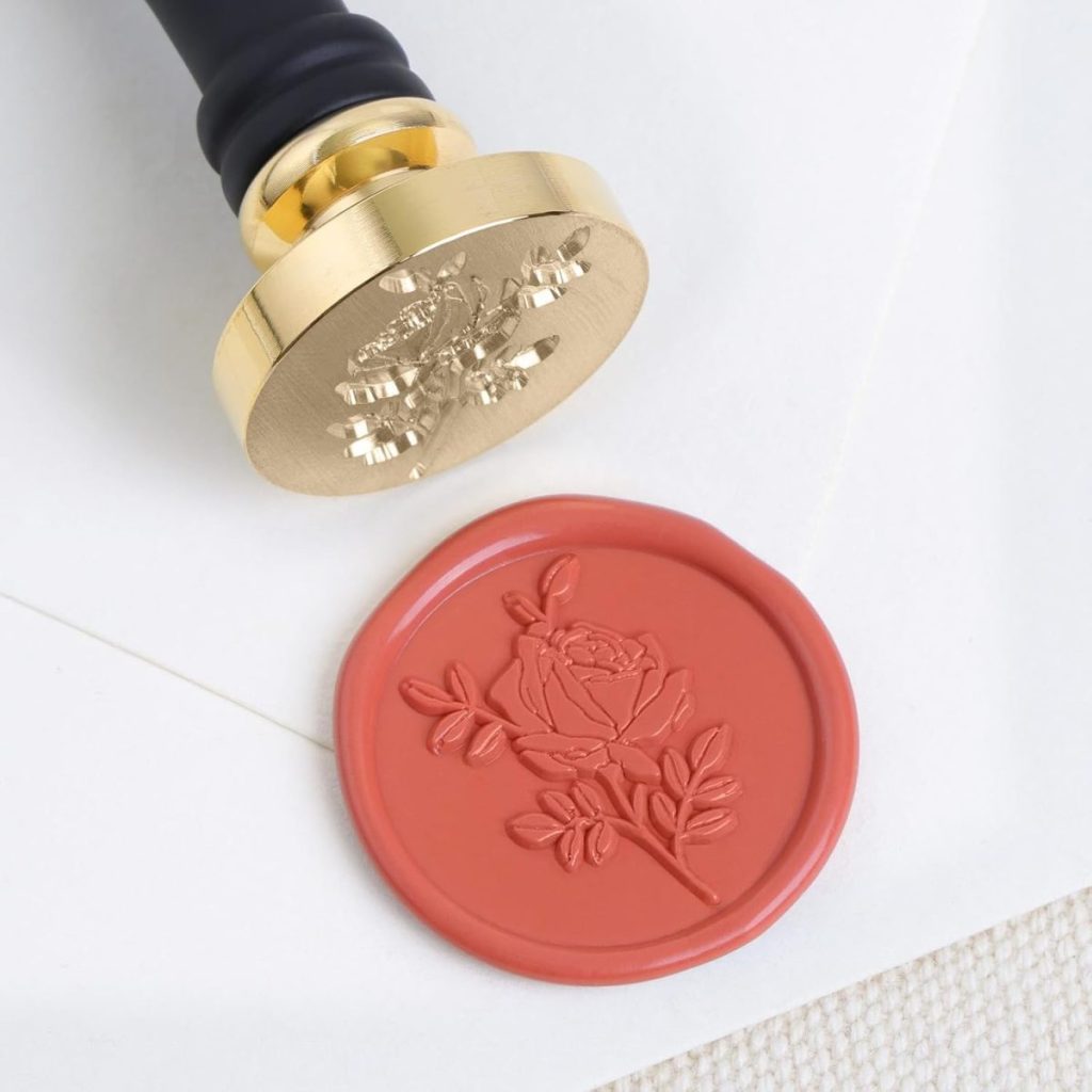 A gold wax seal stamp with a floral design sits beside a red wax seal featuring a rose, showcasing elegant wax seals on a white envelope.