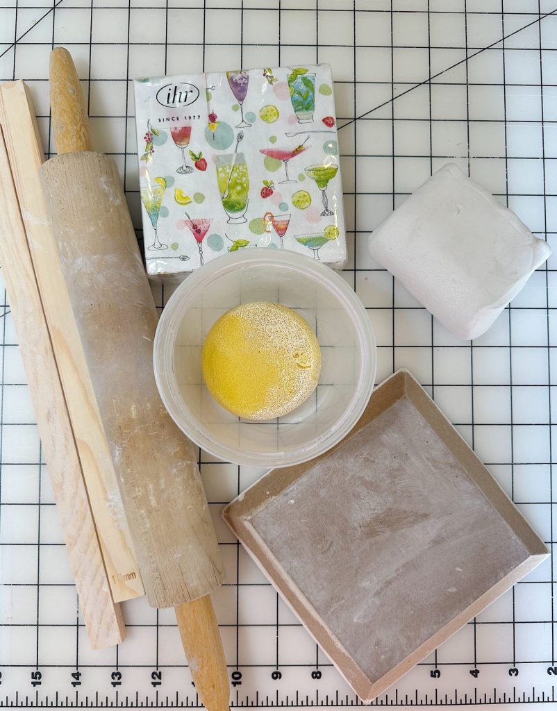A rolling pin, two wooden slats, a napkin, a container with yellow powder, a square slab, and a block of white clay are arranged on a grid cutting mat.