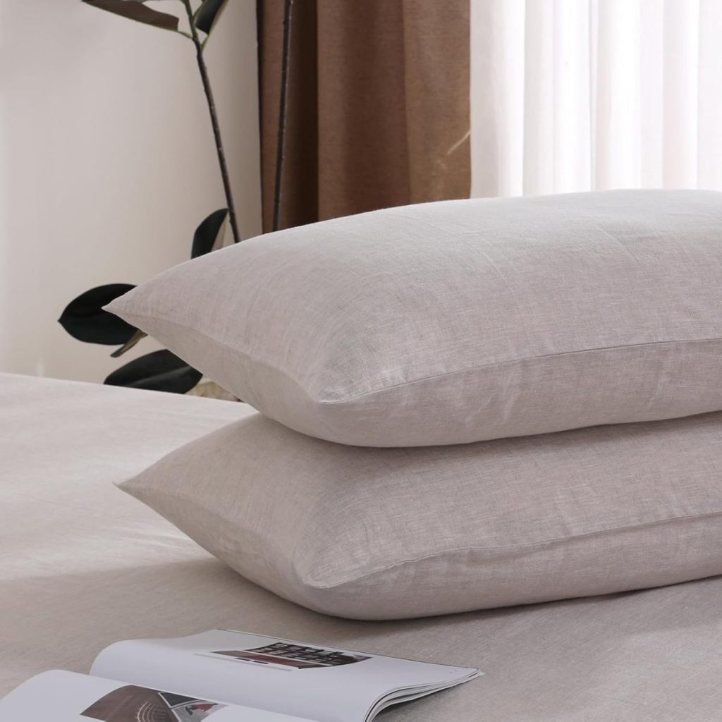 Two beige pillows stacked on a bed with an open magazine nearby; background includes a plant and curtains with natural light.