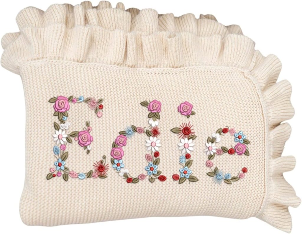 A cream-colored knitted blanket with ruffled edges, featuring the name "Edie" embroidered in colorful floral patterns.