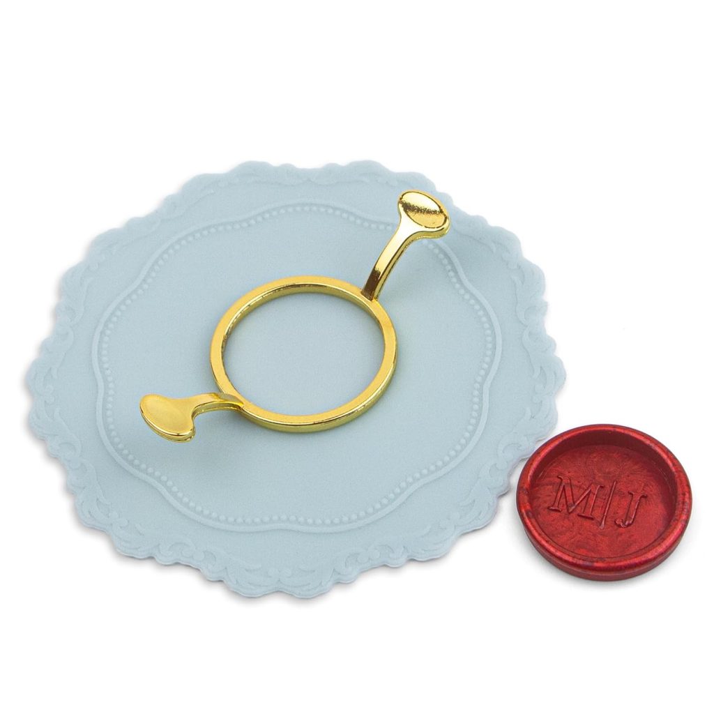 A gold letter opener rests on a decorative light blue paper, beside wax seals including a red one stamped with the initials "MJ".