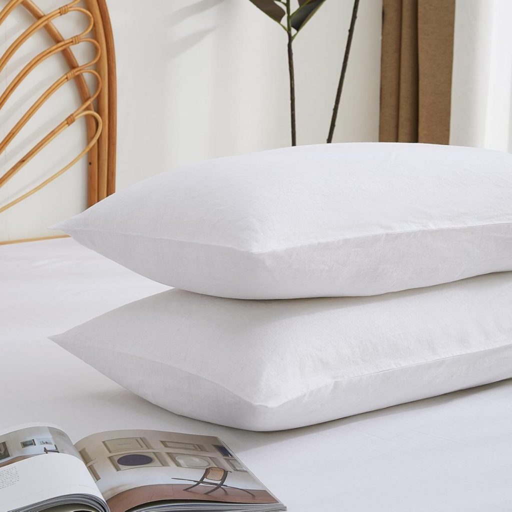 Two white pillows stacked on a white bed with an open magazine nearby; a chair and plant are partially visible in the background.