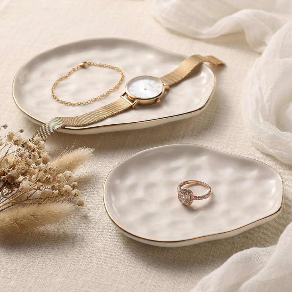 Two ceramic trays with gold rims hold a watch, a bracelet, and a ring. Dried flowers and a white cloth are arranged nearby on a textured light fabric surface.