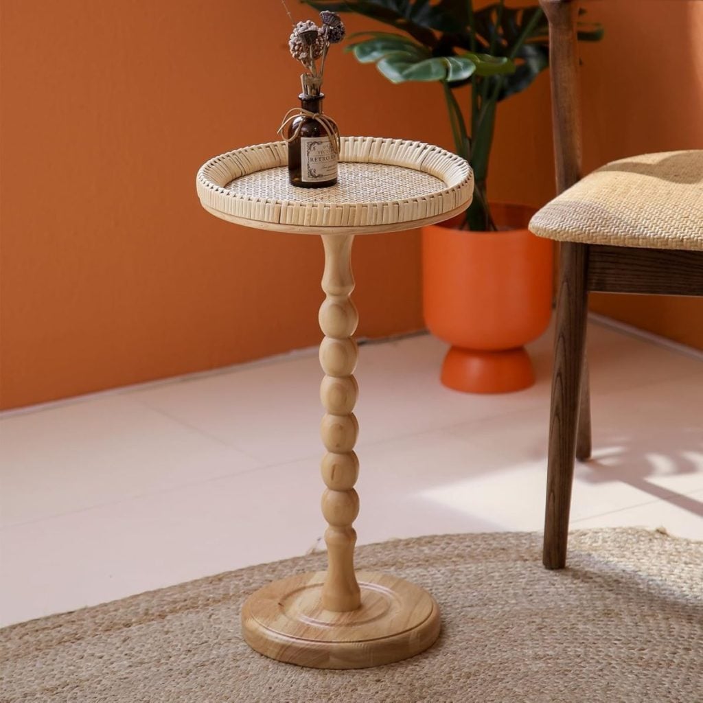 A small wooden side table with a round woven top, holding a brown bottle with dried flowers, placed on a rug near a chair and a plant.