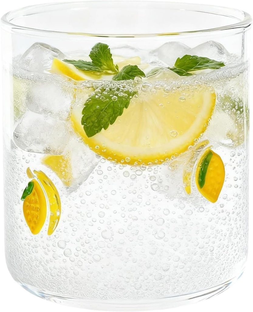 A clear glass of sparkling water with ice, lemon slices, and mint leaves sits atop elegant embroidered cocktail napkins, decorated with small lemon-shaped ornaments on the outside.