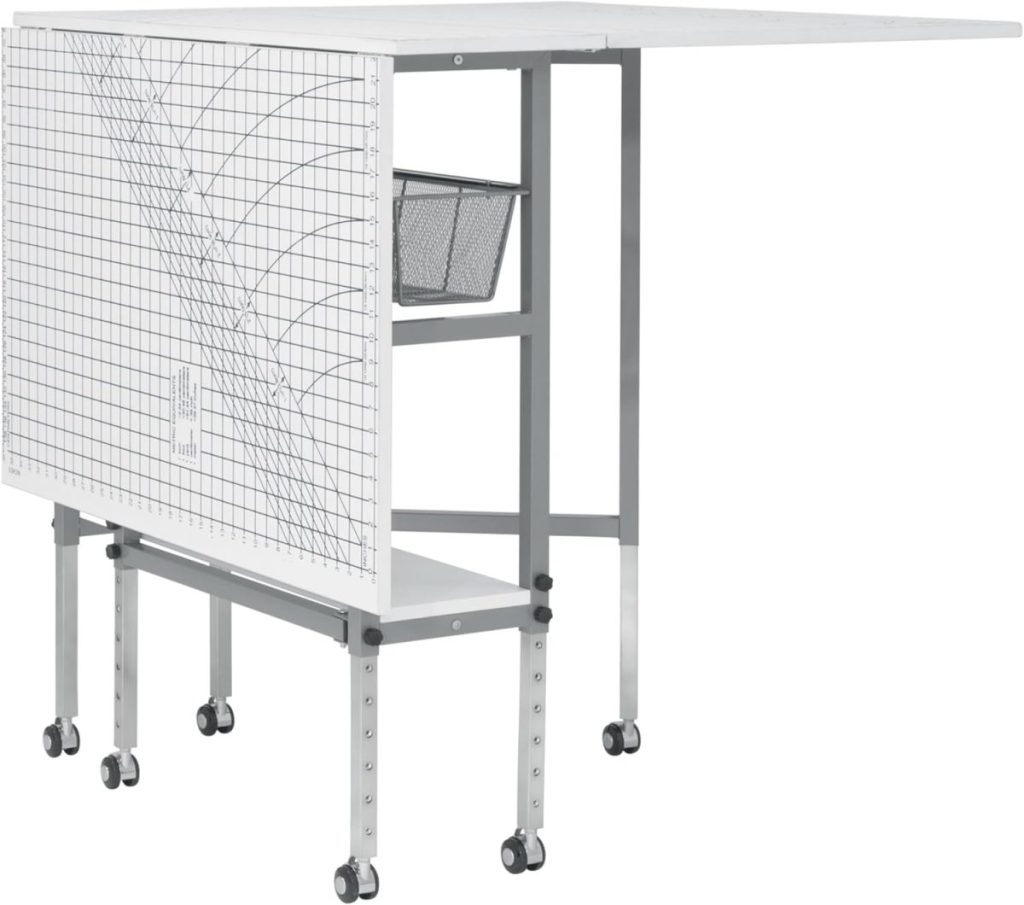 A foldable craft table with a grid marked surface, metal frame, mesh storage basket, lower shelf, and wheels for mobility.