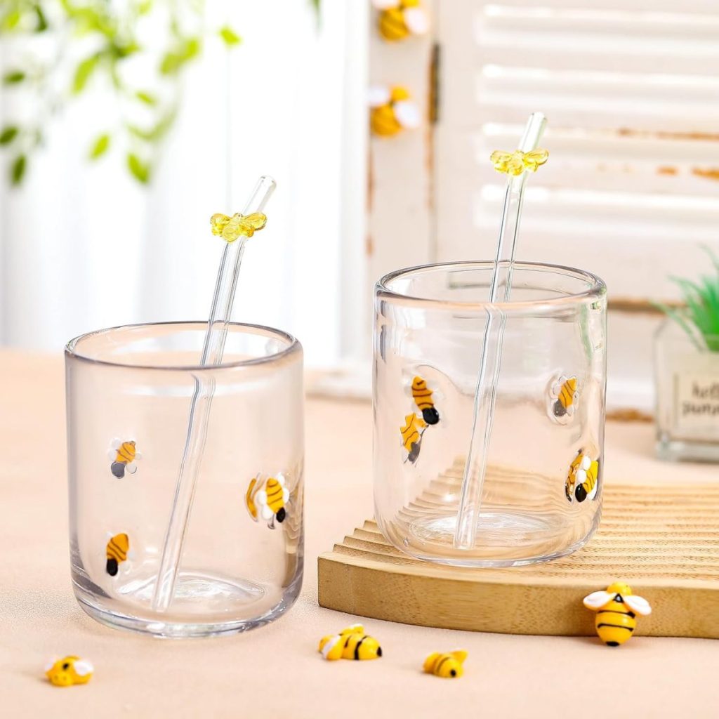 Two clear glasses decorated with bee illustrations, each containing a glass straw with a bee motif at the top, are placed on a table with small bee decorations and elegant embroidered cocktail napkins nearby.