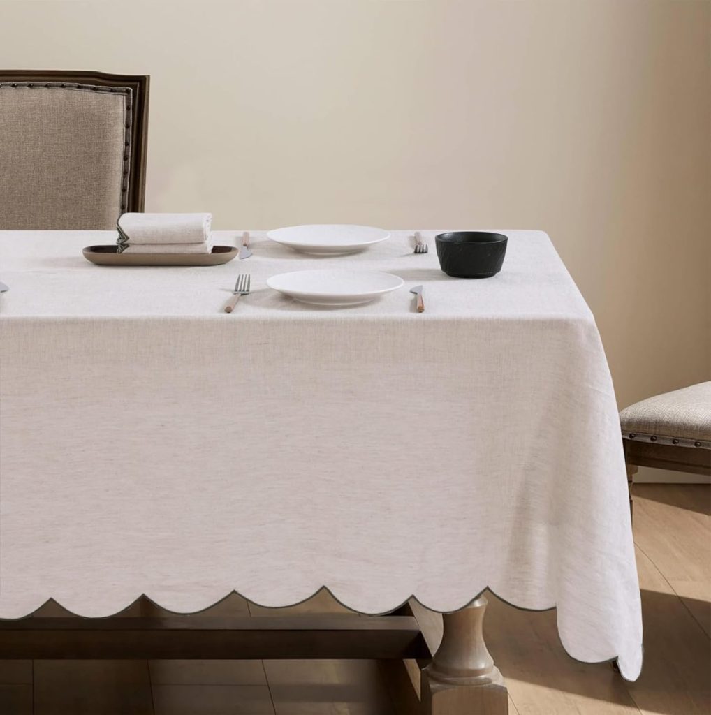A dining table covered with a white scalloped tablecloth is set for two with plates, cutlery, a black bowl, and folded napkins; two upholstered chairs are positioned at the table.