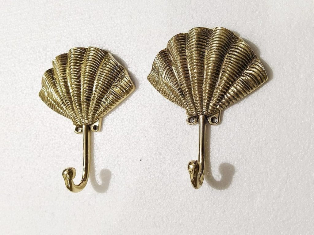 Two brass wall hooks shaped like scallop shells are mounted on a white textured surface, bringing coastal mirror charm to your entryway or bedroom d&eacute;cor.