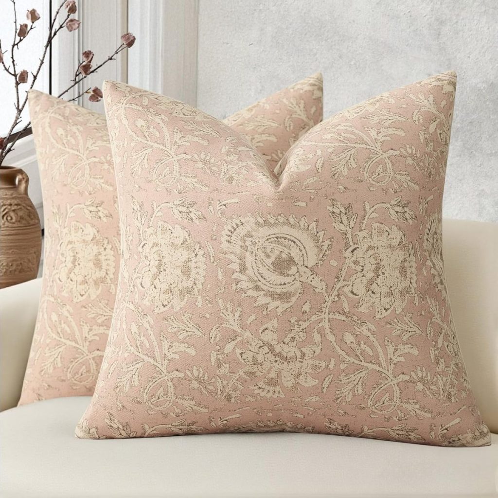 Two square throw pillows with a beige floral pattern on a blush pink background rest on a light-colored sofa, adding a summer crafting touch. A ceramic vase with dried branches sits in the background.