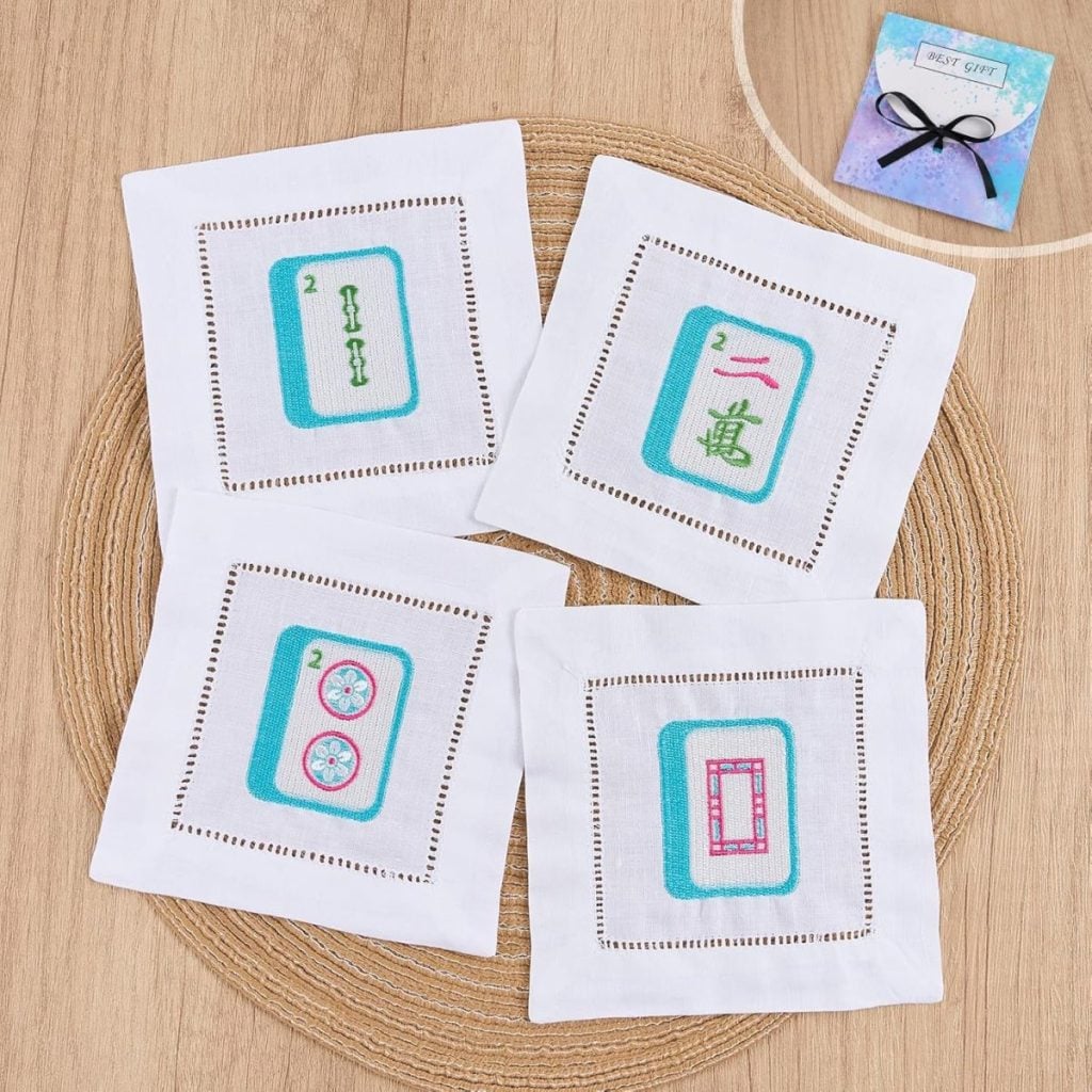 Four white embroidered cocktail napkins featuring mahjong tile designs are arranged on a round mat. A blue gift card with a ribbon is displayed in the top right corner, making this set a thoughtful and stylish gift idea.