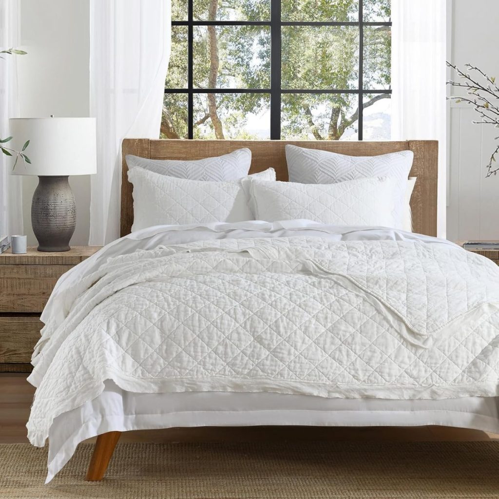 A neatly made bed with white quilted bedding and pillows, a wooden headboard, a bedside lamp, and a window showing trees outside creates a peaceful Memorial Day retreat.