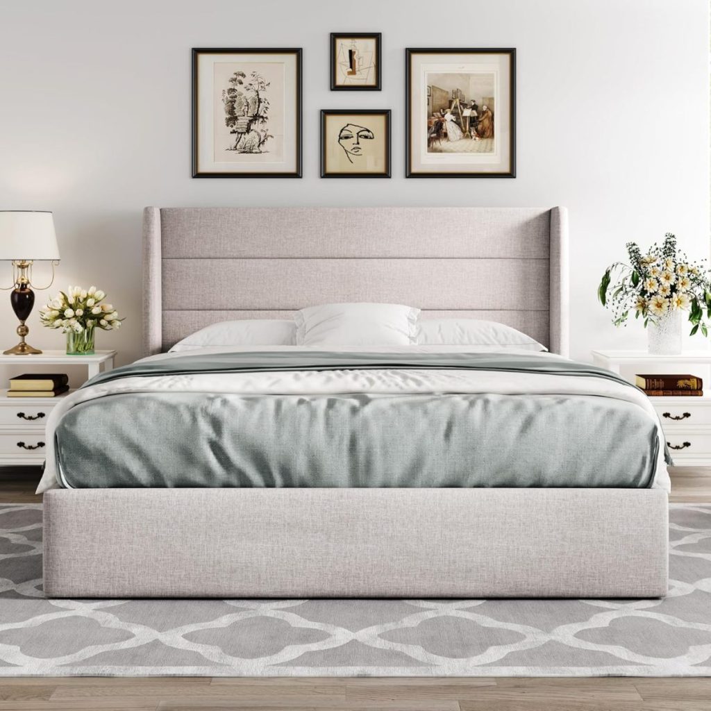A neatly made bed with a light gray upholstered frame, flanked by white nightstands with lamps and flowers, and three framed artworks on the wall above.