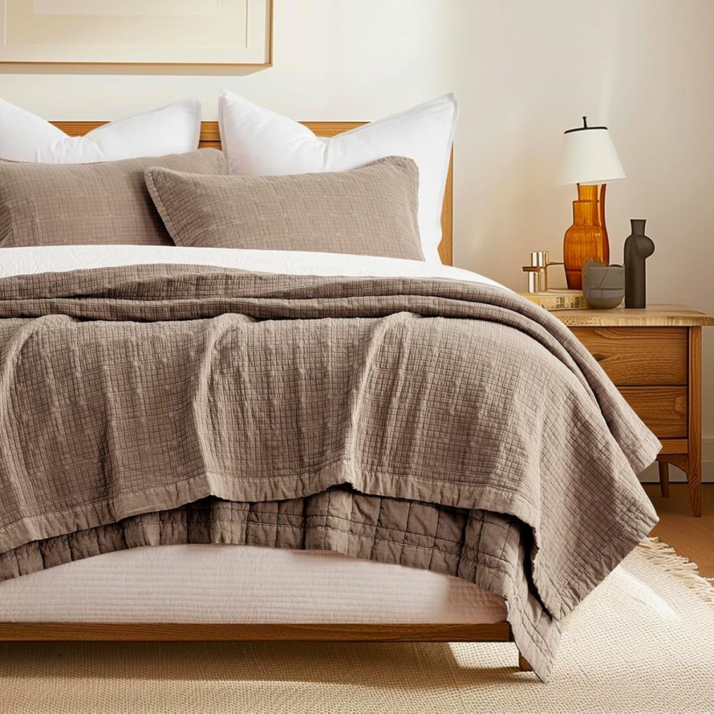 A neatly made bed with taupe bedding and pillows, next to a wooden nightstand holding a lamp, books, and decorative items in a softly lit bedroom.