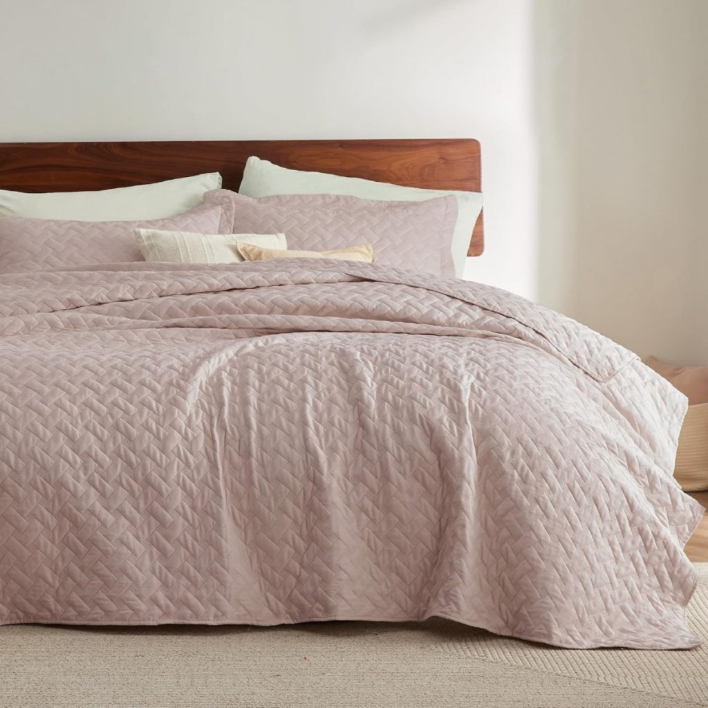 A neatly made bed with a wooden headboard, light pink quilted bedding, and several pillows, set against a plain white wall.
