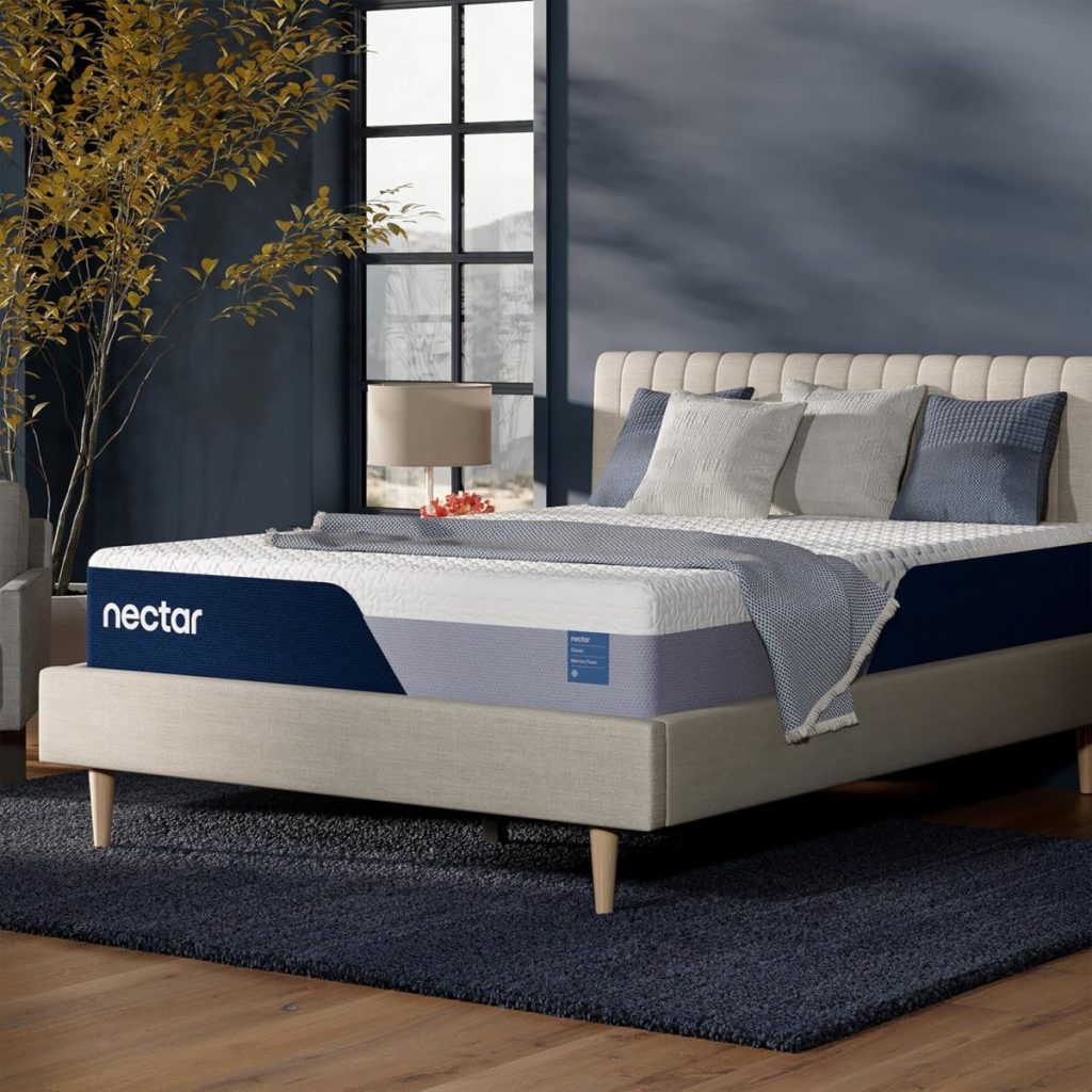 A modern bedroom with a beige upholstered bed, a Nectar mattress, gray bedding, throw pillows, a nightstand with a lamp, and a plant beside a window.