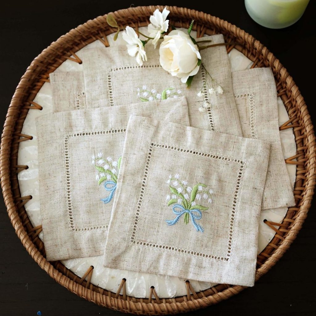 A wicker tray holds embroidered cocktail napkins with floral designs and a blue ribbon, decorated with white artificial flowers on a dark surface.