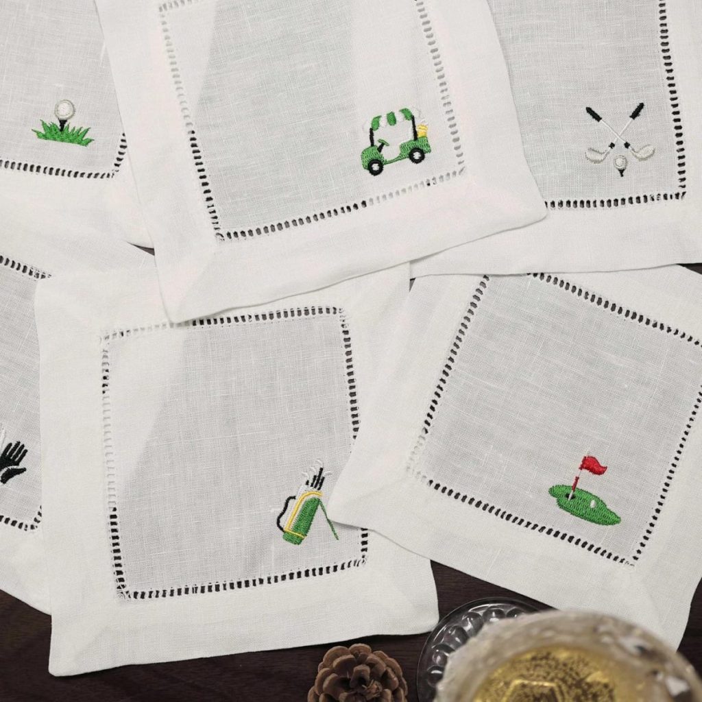 Embroidered Cocktail Napkins featuring golf-themed designs like a cart, clubs, green, and bags are displayed on a dark surface beside a drink and pinecone, adding elegance to any gathering.