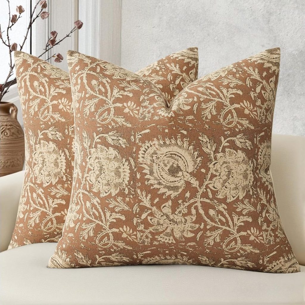 Two decorative throw pillows with a beige floral pattern on a brown background add a touch of summer crafting charm to a light-colored sofa, accented by a vase and branches in the background.