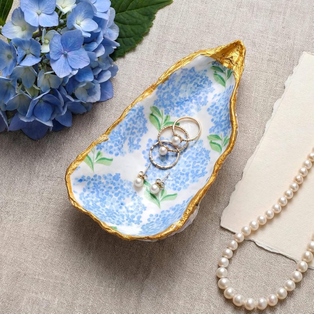 A ceramic dish with blue floral patterns and gold trim holds rings with pearls. A pearl necklace, hydrangea flowers, and paper are nearby on a light fabric surface.