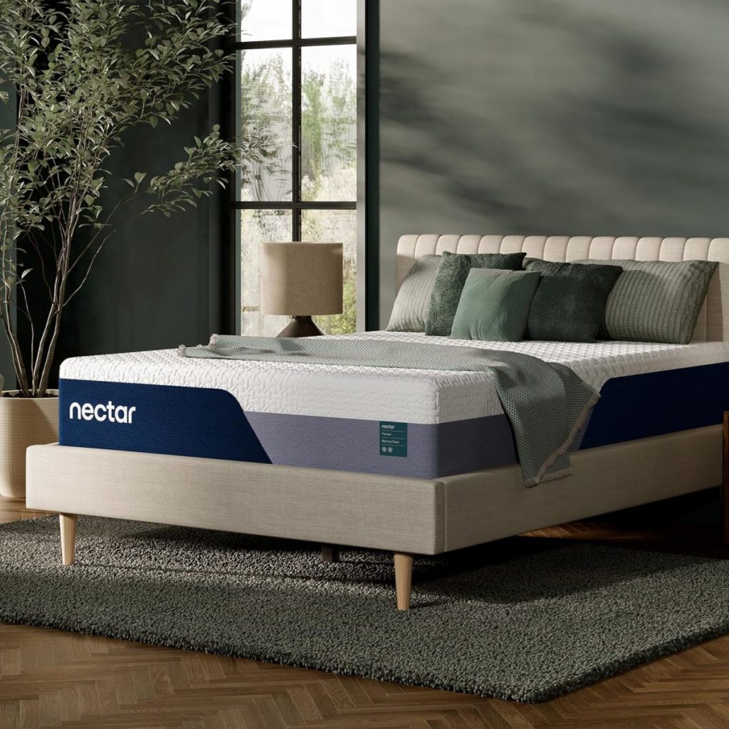 A modern bedroom with a beige upholstered bed frame, a Nectar mattress, green cushions, a lamp, a potted plant, and large windows letting in natural light.