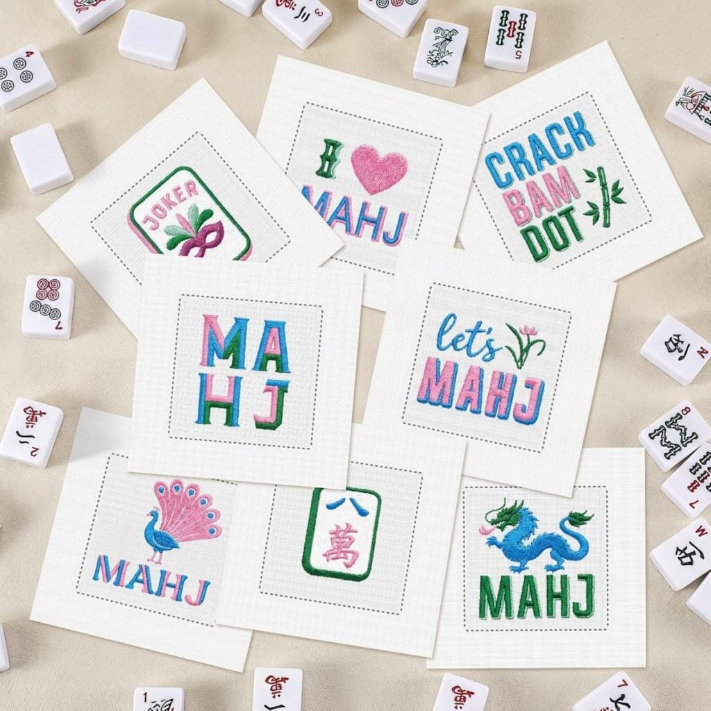 Seven embroidered cocktail napkins featuring cross-stitched Mahjong designs on white cloth are surrounded by Mahjong tiles on a pale surface.