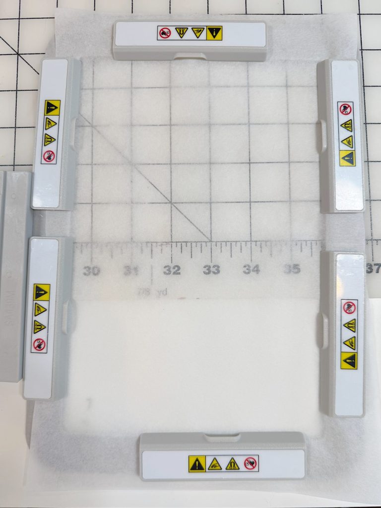 A rectangular plastic frame with multiple warning labels is placed on a gridded cutting mat marked with inch measurements. White padding is visible inside the frame.