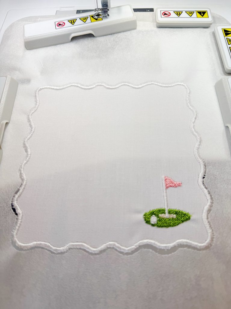 A white fabric square with a stitched wavy border and a small embroidered green golf putting area with a pink flag in the bottom right corner, held in an embroidery machine.