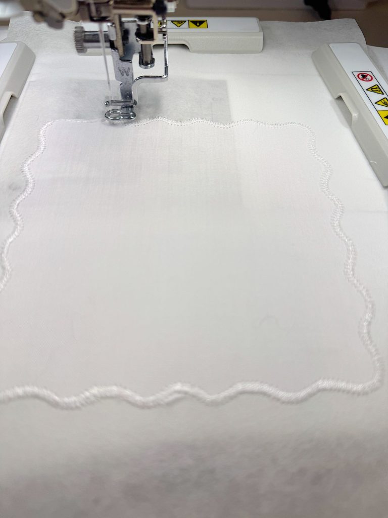 A sewing machine is stitching a scalloped border onto a piece of white fabric secured in an embroidery hoop.