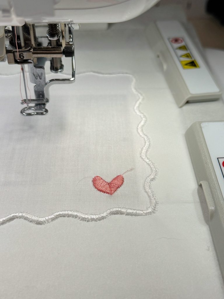 A sewing machine is embroidering a small red heart onto a piece of white fabric with a scalloped edge.