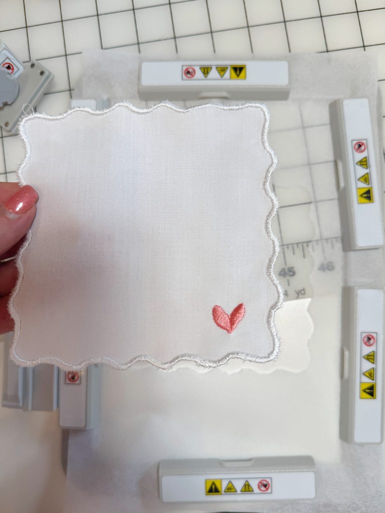 A hand holds a white, scalloped-edge fabric square with a small pink embroidered heart in one corner; sewing tools and a grid mat are visible underneath.