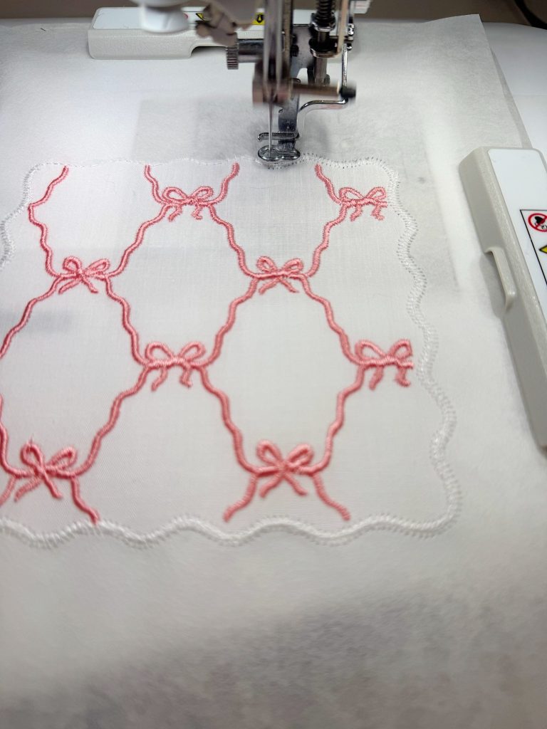 An embroidery machine stitching a white cloth with a pink bow and lattice pattern and a scalloped border.