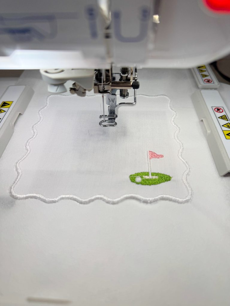 A sewing machine is embroidering a scalloped-edge white cloth with a small green golf course and a red flag design in the corner.