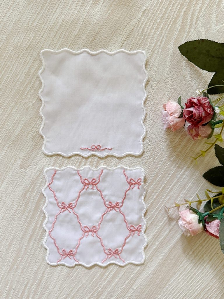 Two white handkerchiefs with scalloped edges; one plain and one embroidered with a pink ribbon lattice pattern, placed beside pink artificial flowers on a light wooden surface.