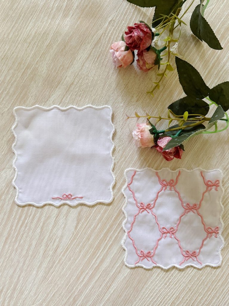 Two white handkerchiefs with pink embroidery are displayed on a light wood surface next to artificial pink roses and green leaves.